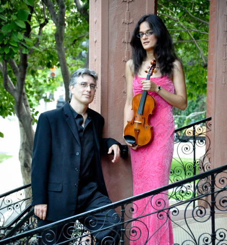 Photo of Olivia Hajioff and Marc Ramirez, standing outside on a staircase. He is on
		the left in a dark blue suit with an open collar, leaning on his left arm and looking at the camera. She is next
		to him in a long fuschia gown, holding a violin and looking at the camera.